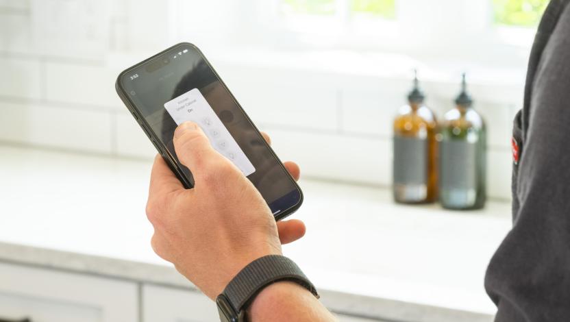 Person adjusting smart kitchen lighting using a mobile app on a smartphone near a white countertop and backsplash