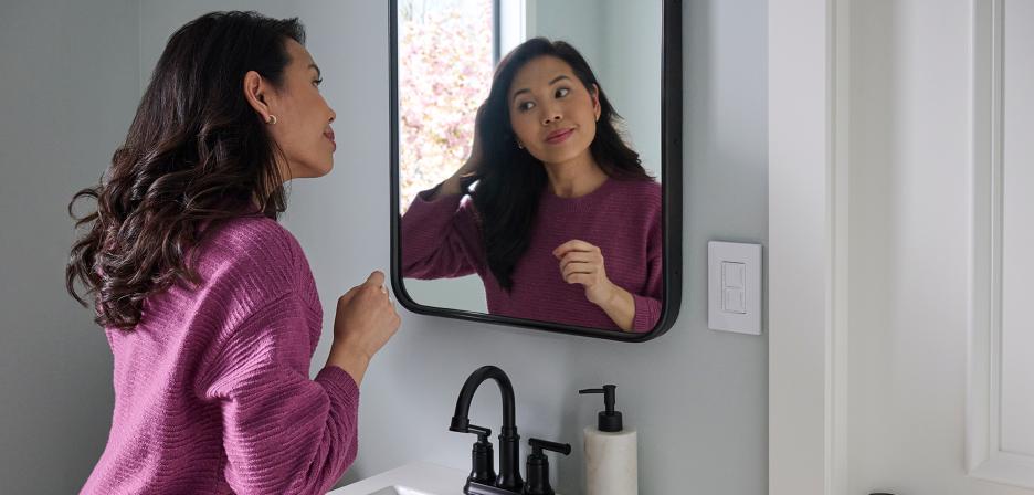 Woman styling her hair in a small bathroom, with a Dual Dimmer and Timer Switch visible on the wall nearby.