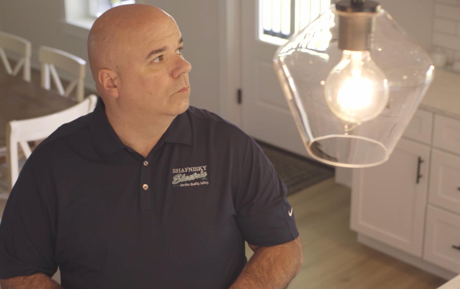 Electrician in a navy Shafnisky Electric shirt looking up at a lit pendant light in a modern kitchen