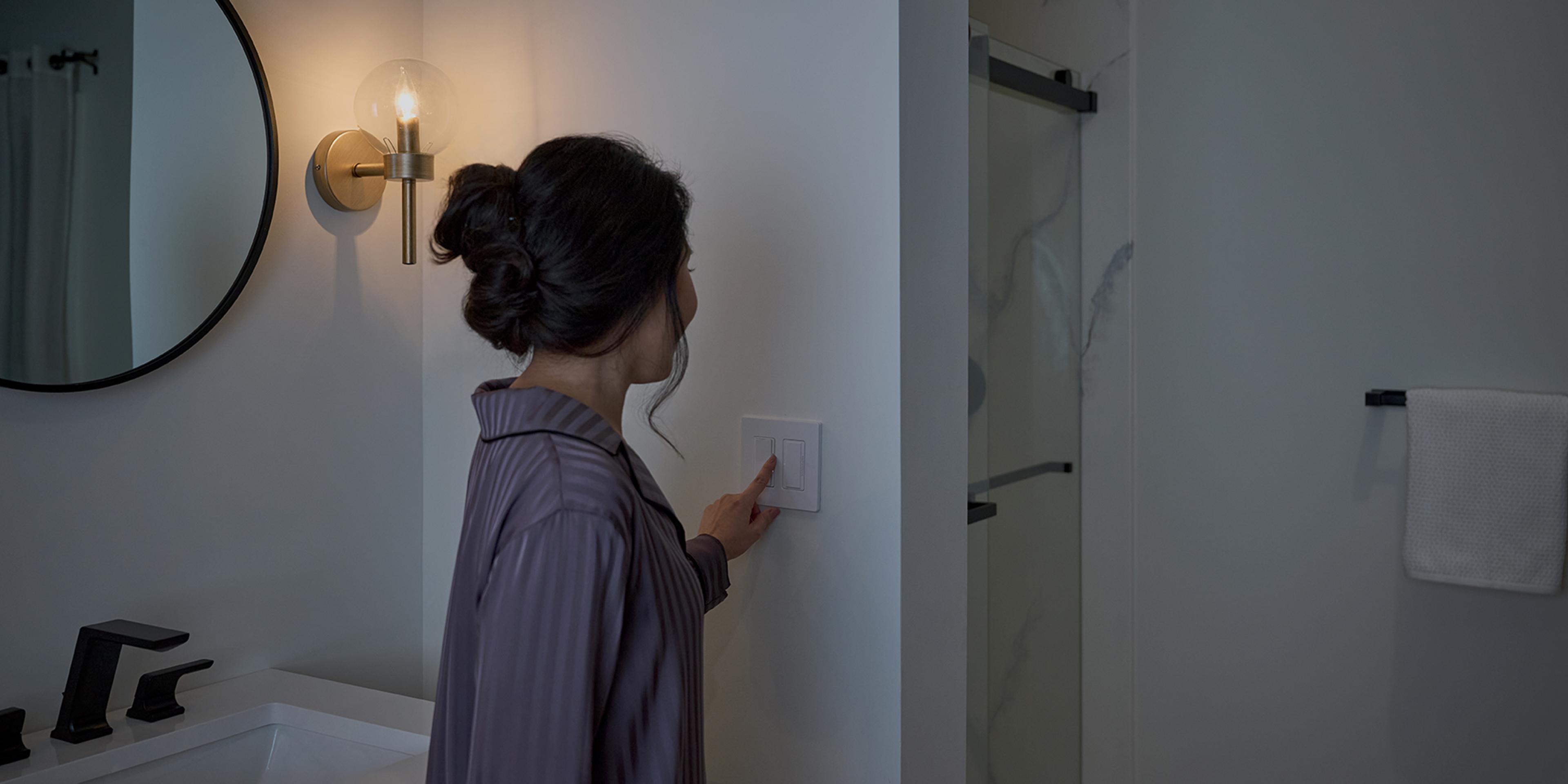 Woman adjusting a Diva dimmer with Night Mode, ganged with a Humidity Sensor switch, in a bathroom setting.